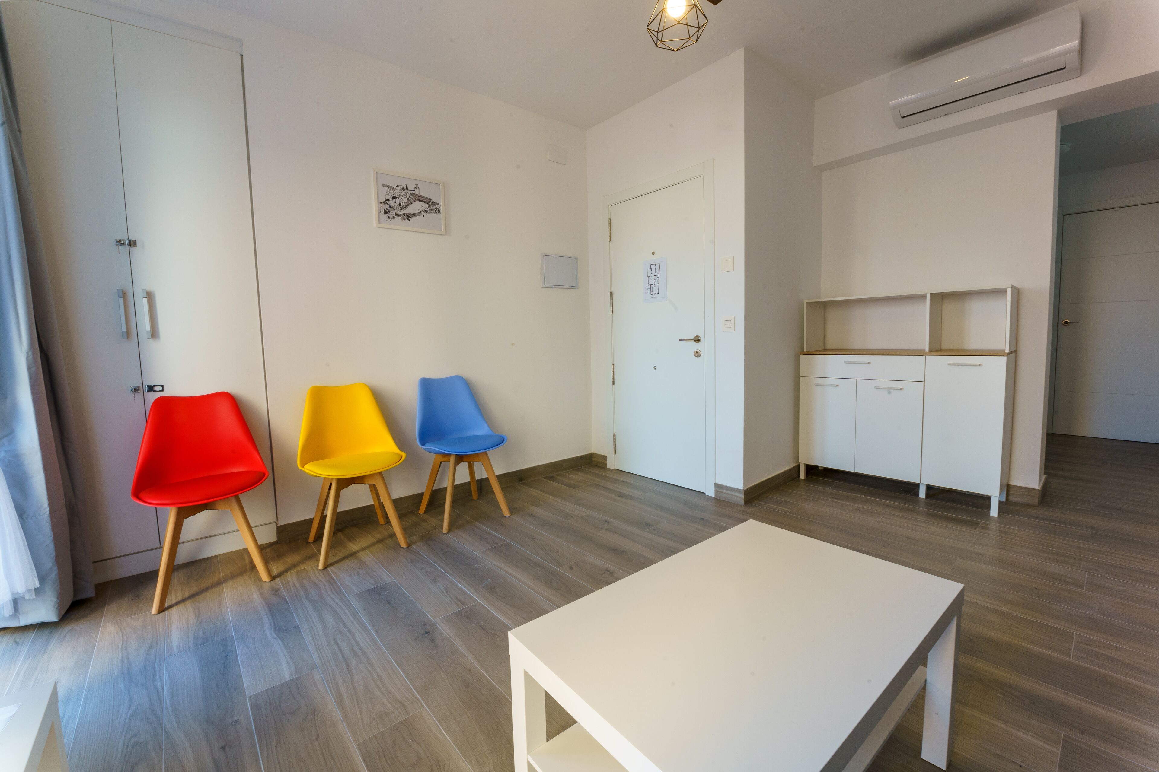 Ein Zimmer mit einem weißen Tisch, drei Stühlen in Rot, Gelb und Blau sowie einem weißen Kleiderschrank.