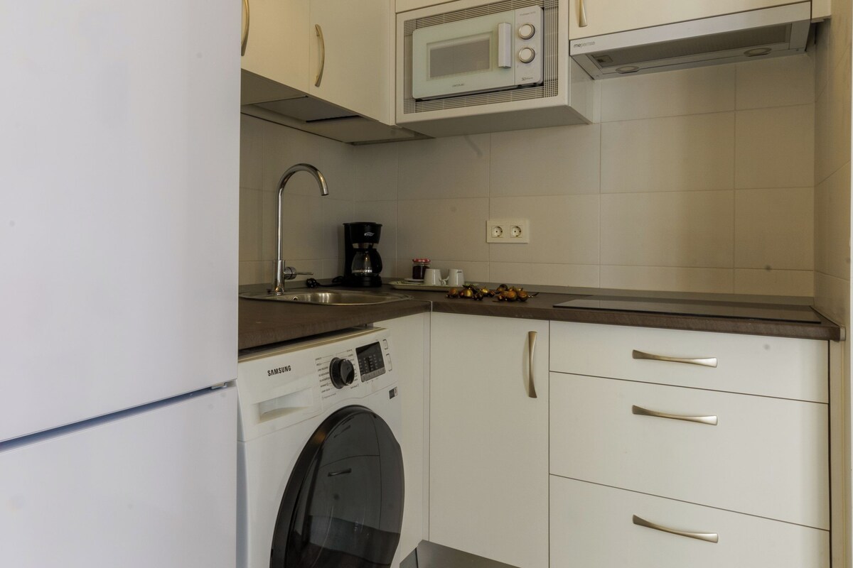 Una cocina compacta con gabinetes blancos, microondas integrado, lavadora y cafetera.
