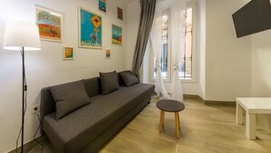 A modern living room with a grey sofa, a small round table, and a white bench. There are wall art pieces and a lamp on the left. - APARTAMENTOS MAPAMUNDI (Badajoz)