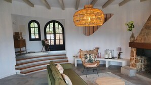 Ett rymligt vardagsrum med en grön soffa, en fåtölj i rotting, ett soffbord i trä, en eldstad i sten och en stor, vävd lampa.