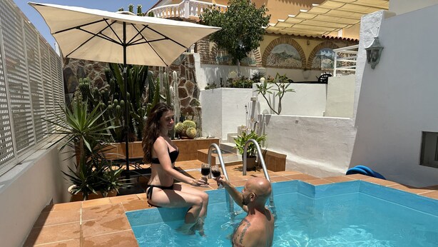 En swimmingpool med flisebelagt område, stenvæg og en have med kaktusser.