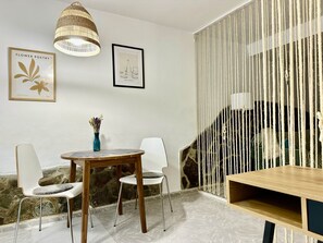 Une pièce moderne avec une table ronde en bois, deux chaises blanches, un vase bleu contenant des fleurs séchées, un luminaire en fibres tressées et un tableau encadré accroché au mur.