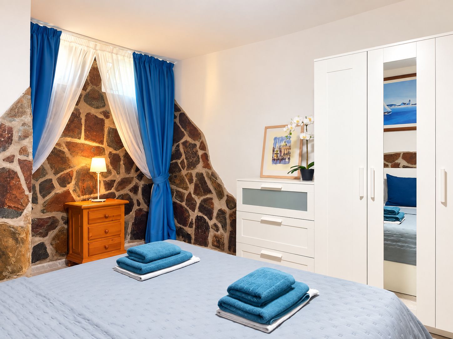Une chambre avec un mur en pierre, un lit avec des serviettes bleues, une table de chevet avec une lampe et une armoire ornée d’un tableau.
