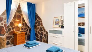 Une chambre avec un mur en pierre, un lit avec des serviettes bleues, une table de chevet avec une lampe et une armoire ornée d’un tableau.