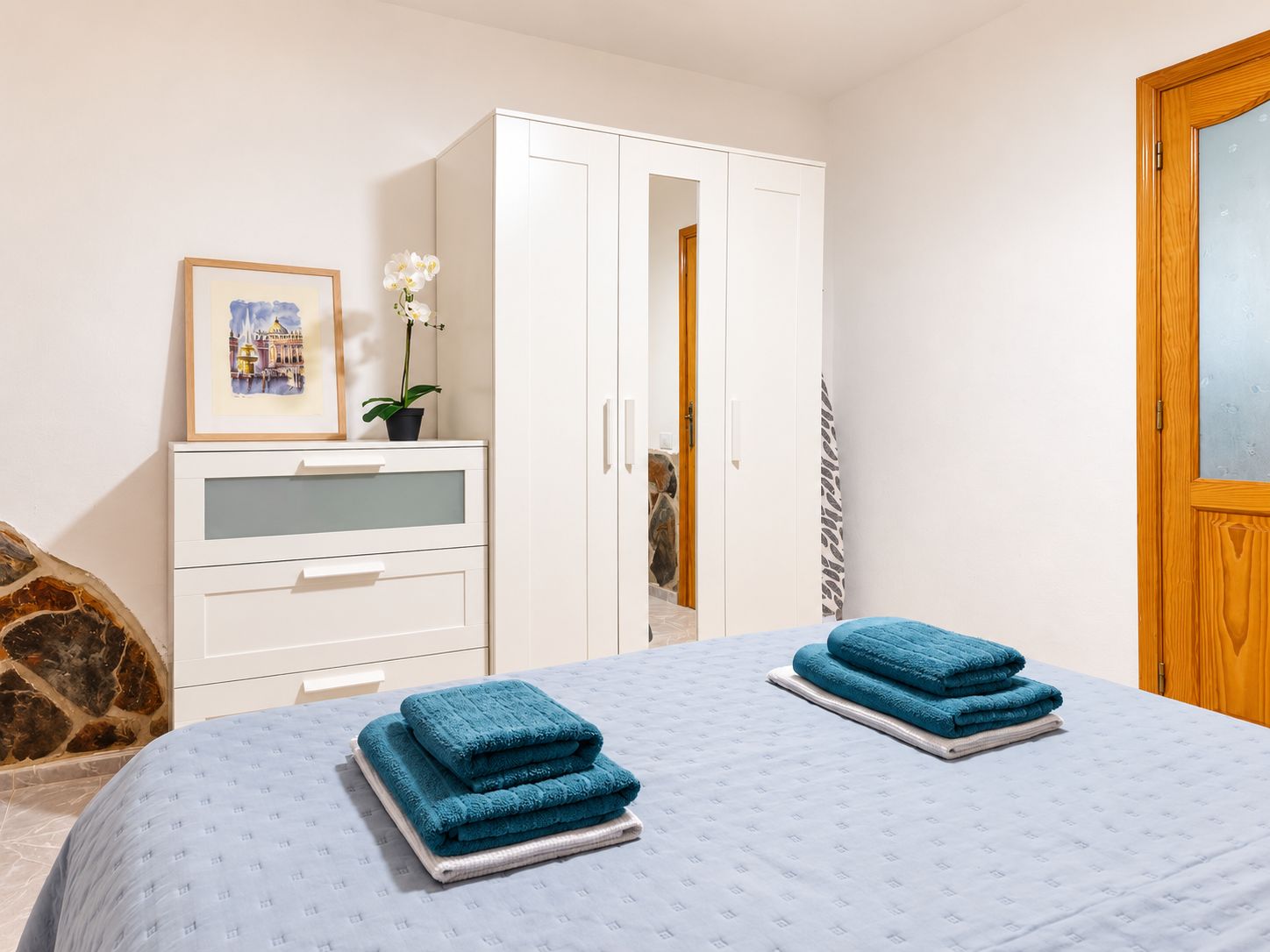 Ein Schlafzimmer mit Bett, Kleiderschrank, Schminktisch, einem gerahmten Bild und einer Zimmerpflanze.