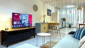 En moderne stue med et fladskærms-tv, en sofa, et lille rundt bord og et spiseområde med en stol og en lampe.