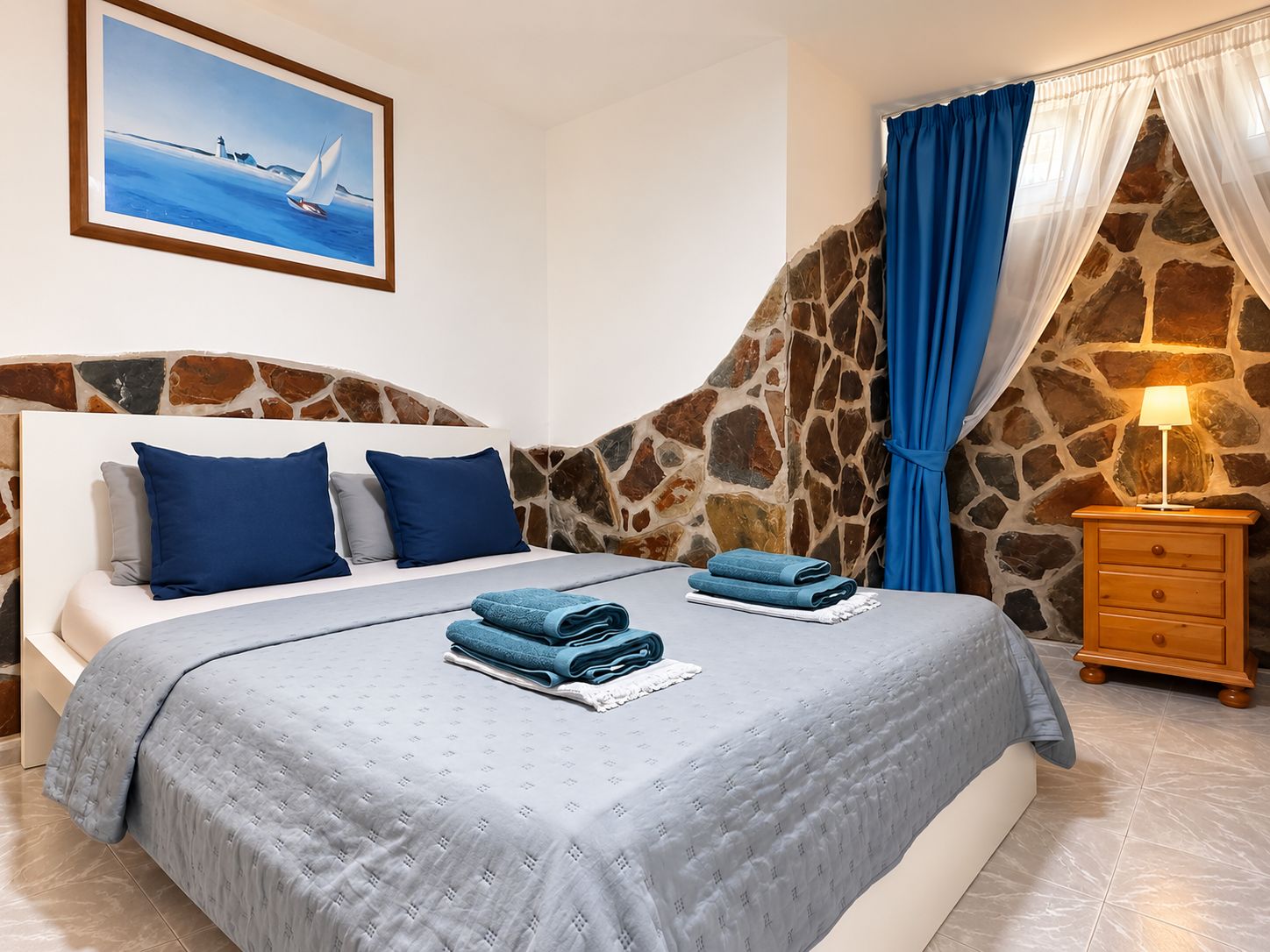 A bedroom with a stone wall, a bed with blue bedding, a nightstand with a lamp, and a painting of a sailboat.