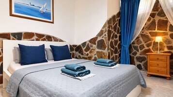A bedroom with a stone wall, a bed with blue bedding, a nightstand with a lamp, and a painting of a sailboat.
