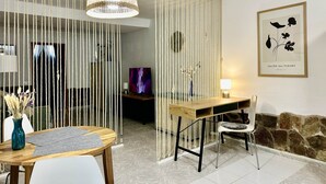 Et værelse med et træspisebord, hvide stole, en hængende vævet lampe, et fjernsyn og et indrammet botanisk tryk på væggen.