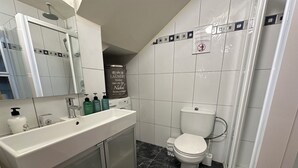 Un baño con lavamanos blanco, espejo e inodoro. Hay artículos de aseo en el lavamanos y un cabezal de ducha sobre el espejo.