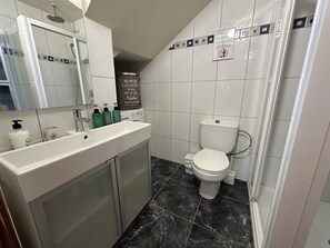Un baño con lavamanos blanco, espejo e inodoro. Hay artículos de aseo en el lavamanos y un cabezal de ducha sobre el espejo.