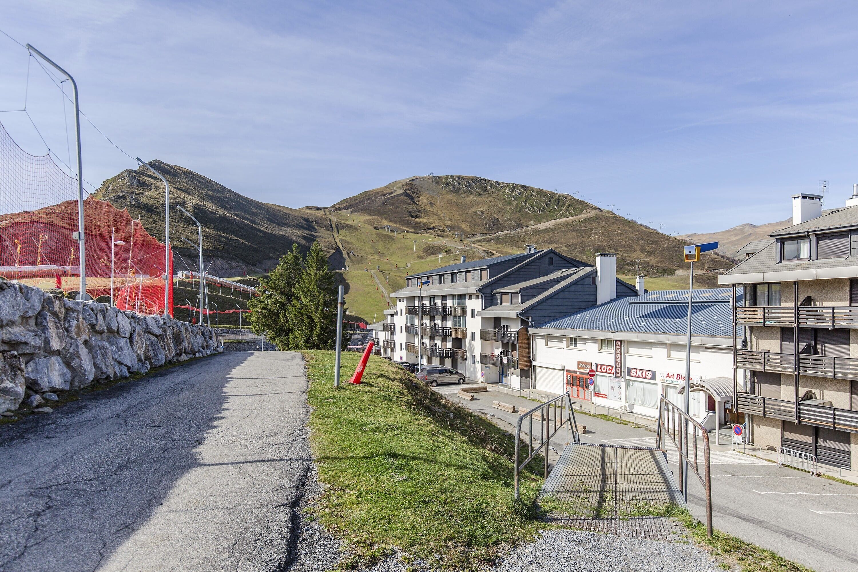 Une route de montagne bordée d’un muret en pierre, un cône de signalisation rouge et un bâtiment doté de balcons et d’une enseigne sur laquelle on peut lire « LOCA SKIS ».