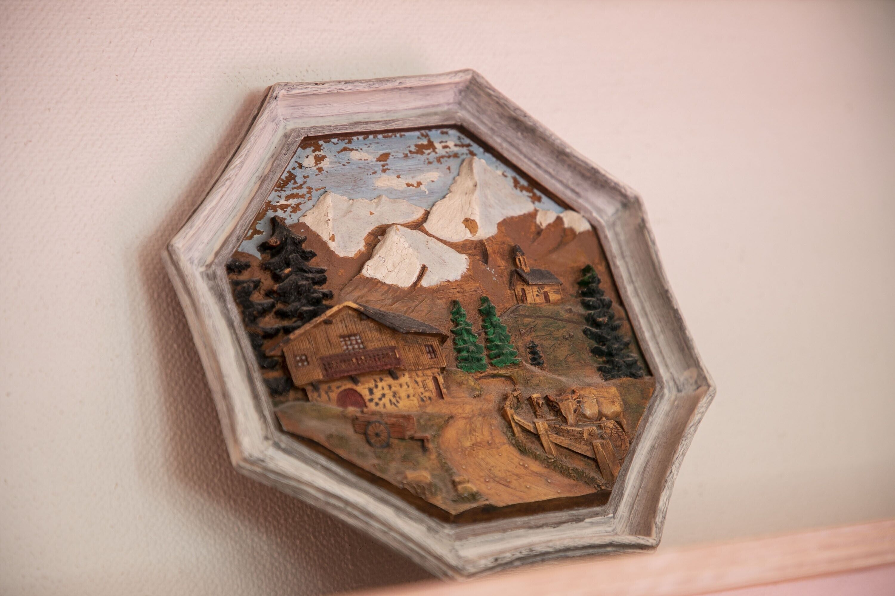 A hexagonal wooden frame with a rustic scene including a house, mountains, and trees.