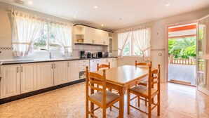 Een keuken met witte kastjes, een eettafel met stoelen en uitzicht op groen door de ramen.