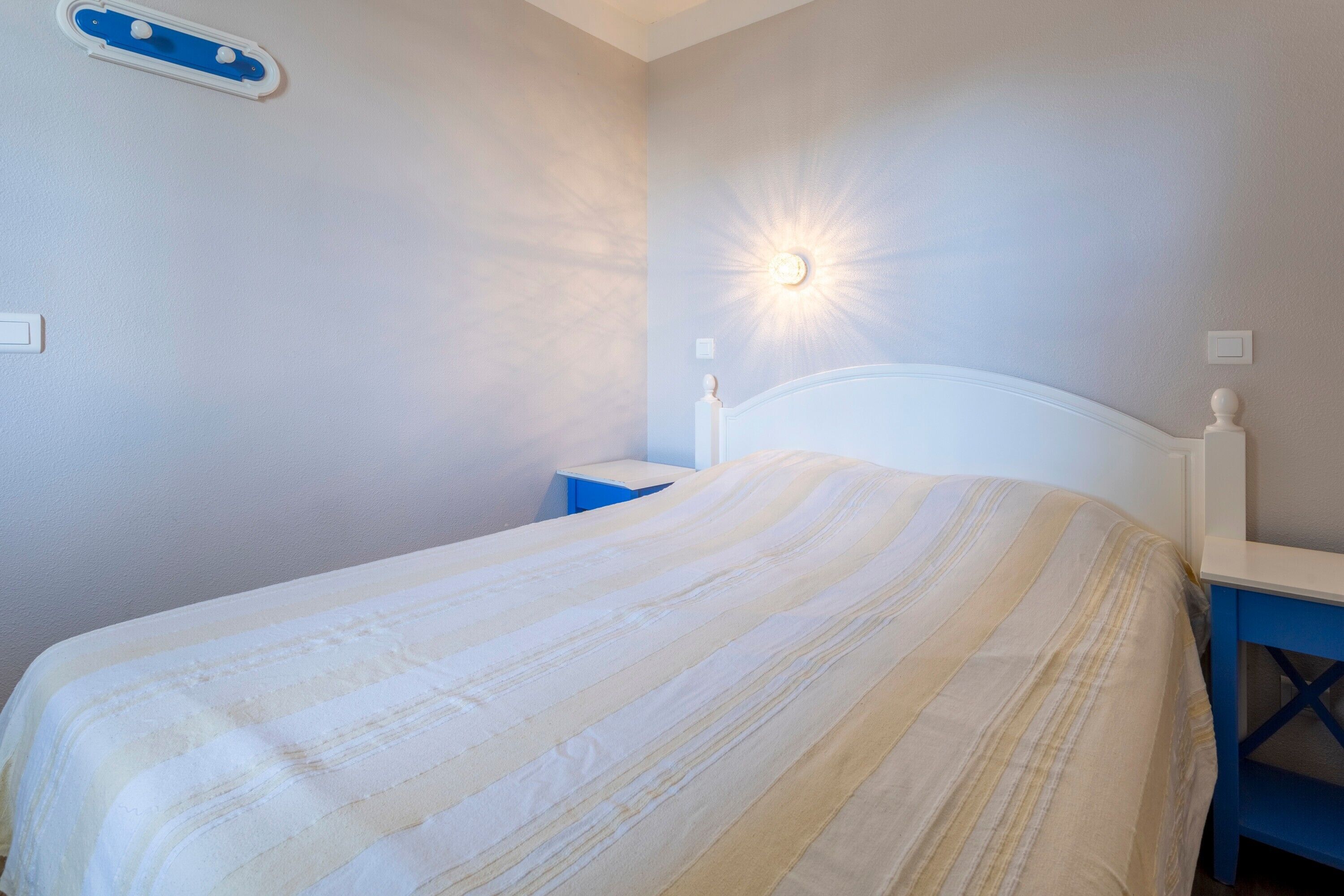 Een net opgemaakt bed met een gestreept dekbed, een nachtkastje en een wandlamp.