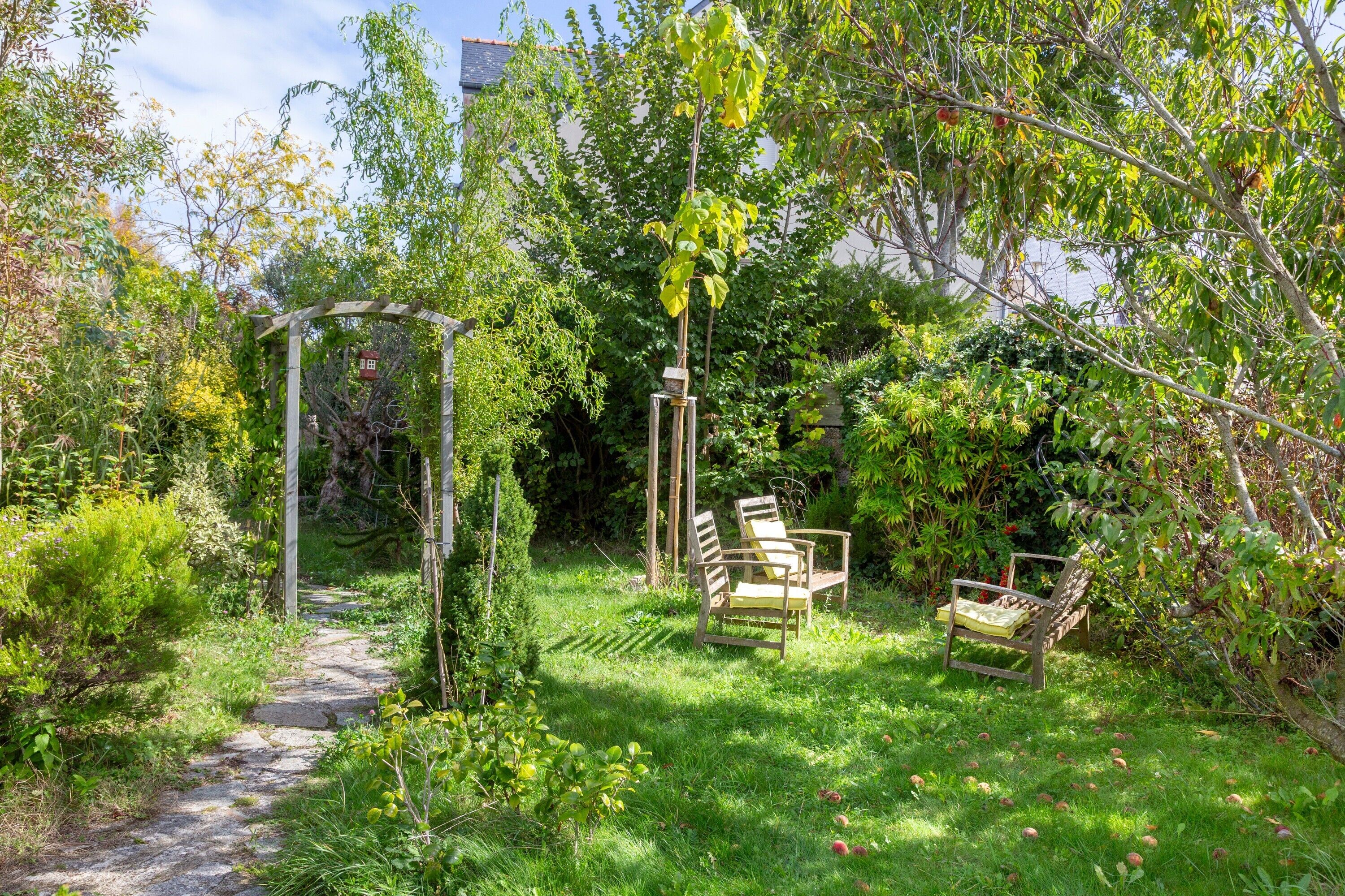 Un jardin avec un chemin de pierres, une pergola en bois et deux chaises en bois.