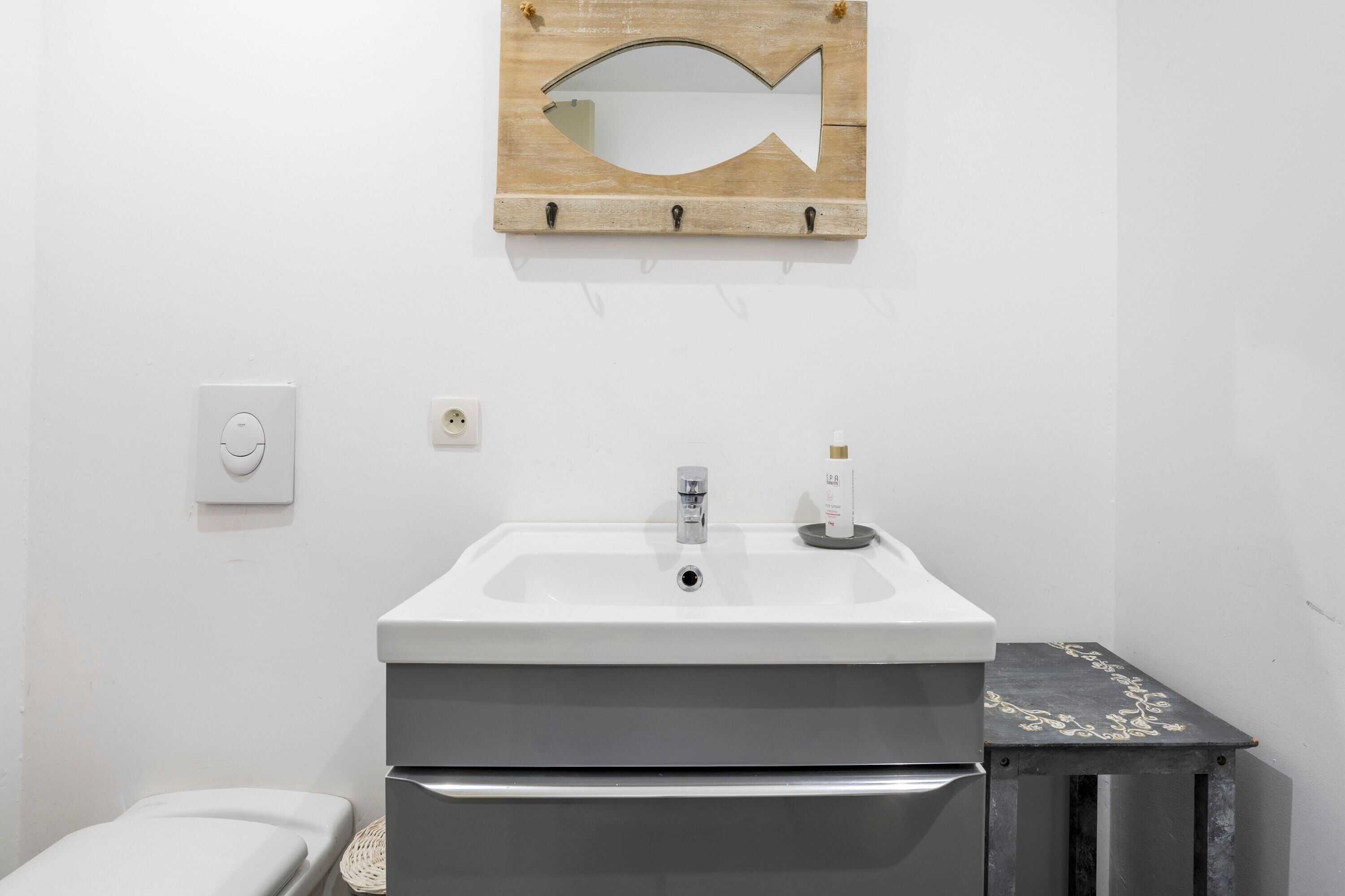 Een badkamer met een witte wastafel, een spiegel in de vorm van een houten vis, en een klein zwart kastje.