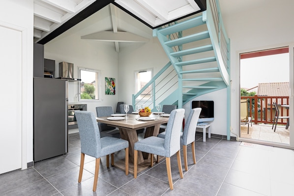 A modern kitchen with a built-in refrigerator, a dining area with a wooden table and grey upholstered chairs, and a blue spiral staircase leading to an upper level.