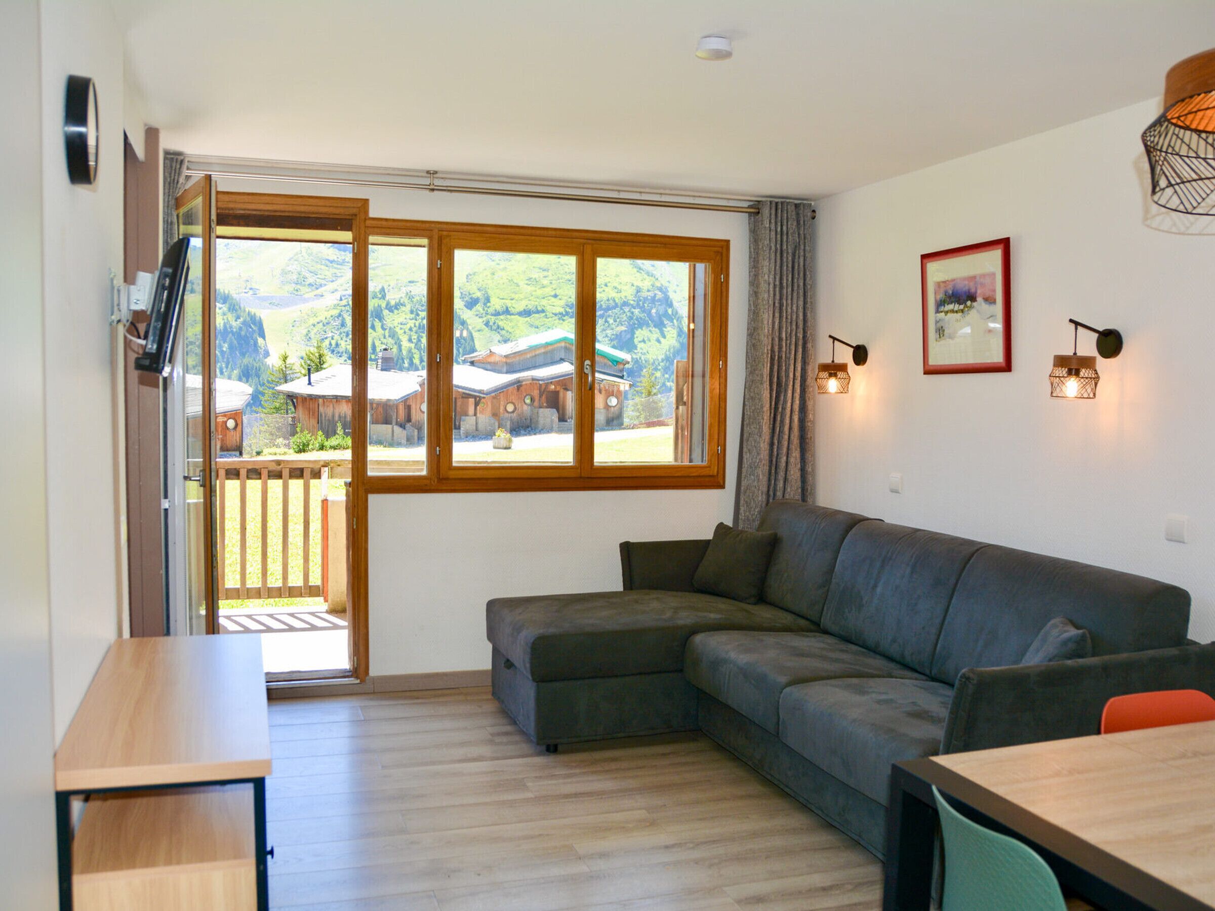 A living room with a wooden floor, a grey sofa, a wooden table, and a large window with a view of buildings and greenery.