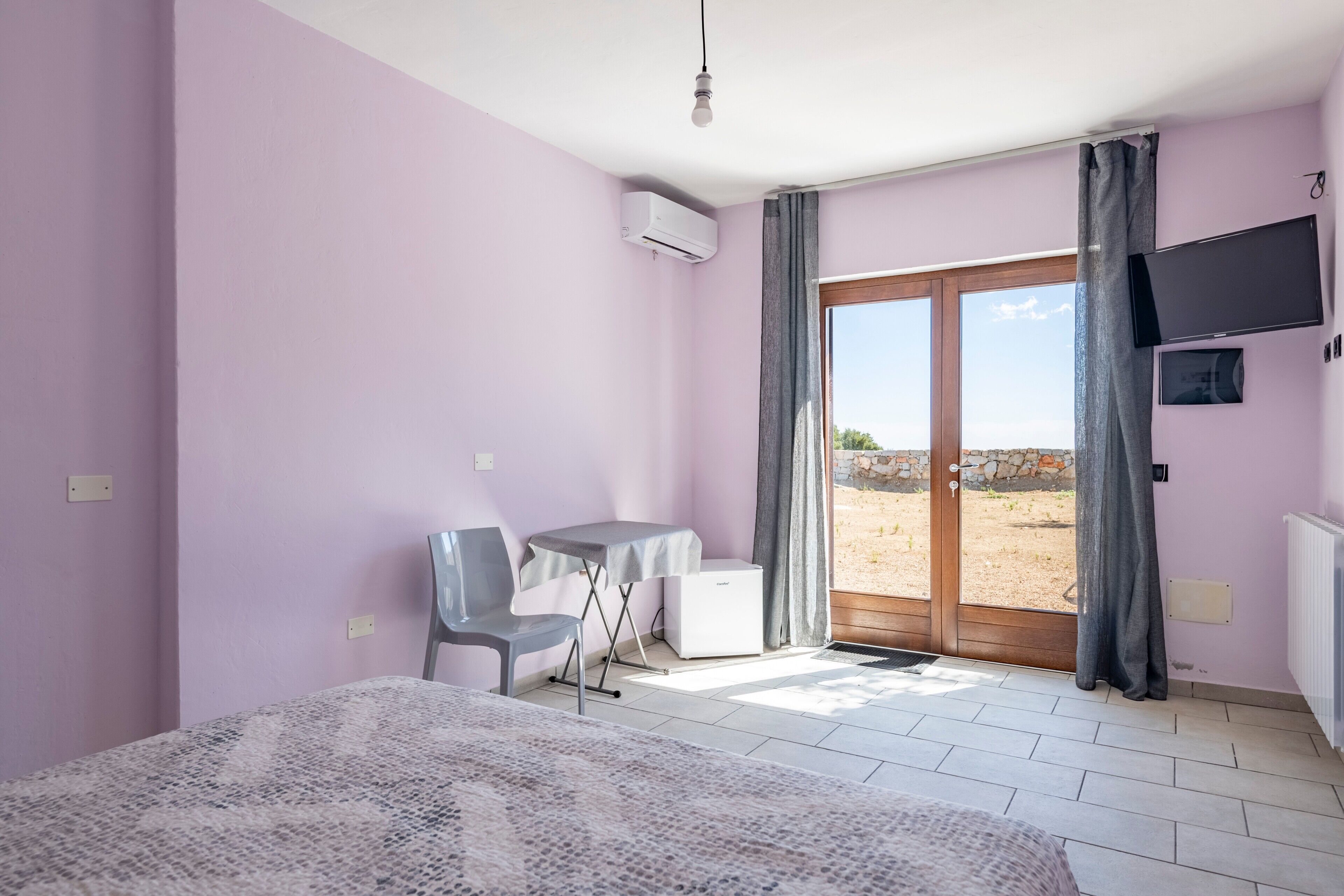 Una habitación con cama, escritorio, silla, televisor y puerta corrediza de vidrio a un espacio exterior.