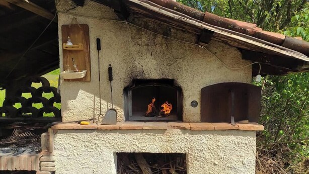 Un four en briques rustique installé à l’extérieur, avec un feu qui crépite à l’intérieur, entouré d’une structure en bois et de verdure.