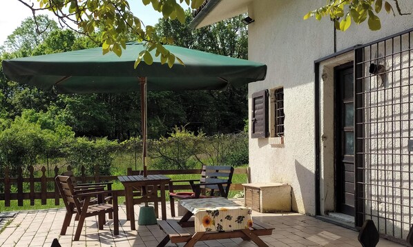 Une terrasse extérieure avec des meubles en bois, un parasol vert et un bâtiment blanc doté d’une fenêtre grillagée.