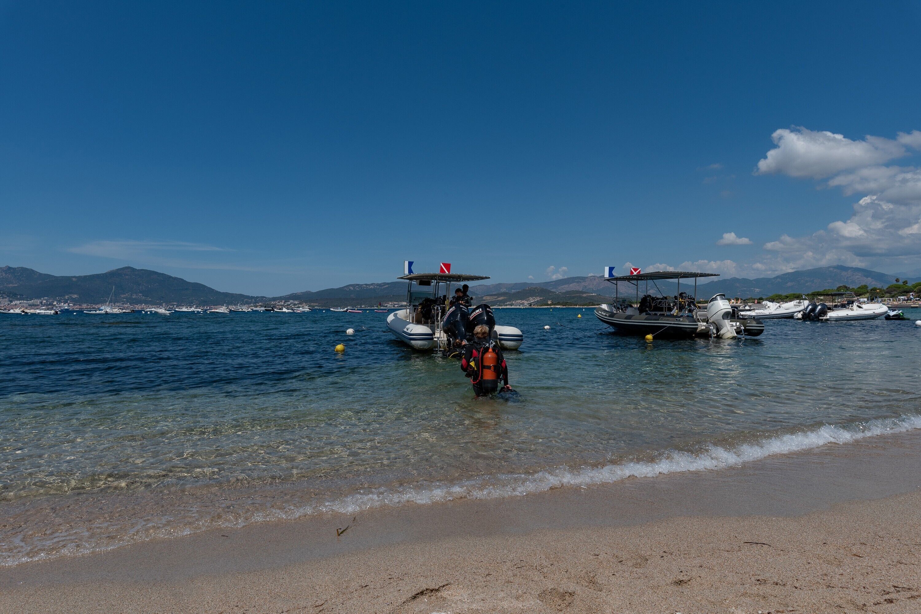 Una platja amb vaixells, aigua cristal·lina i muntanyes a la distància.