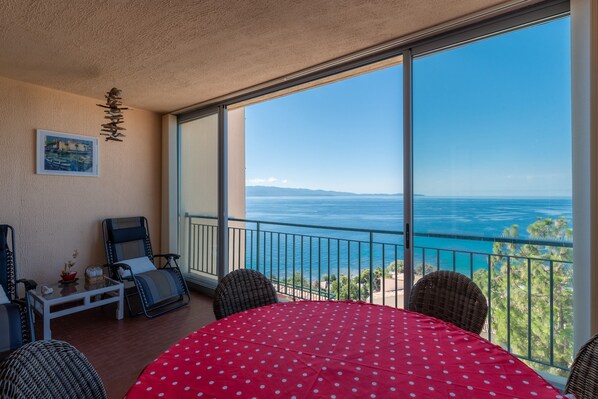 A balcony with a view of the ocean, a table with a red polka dot tablecloth, and wicker chairs.