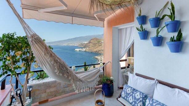 A balcony with a hammock, potted plants, a wicker basket, and a view of the sea.