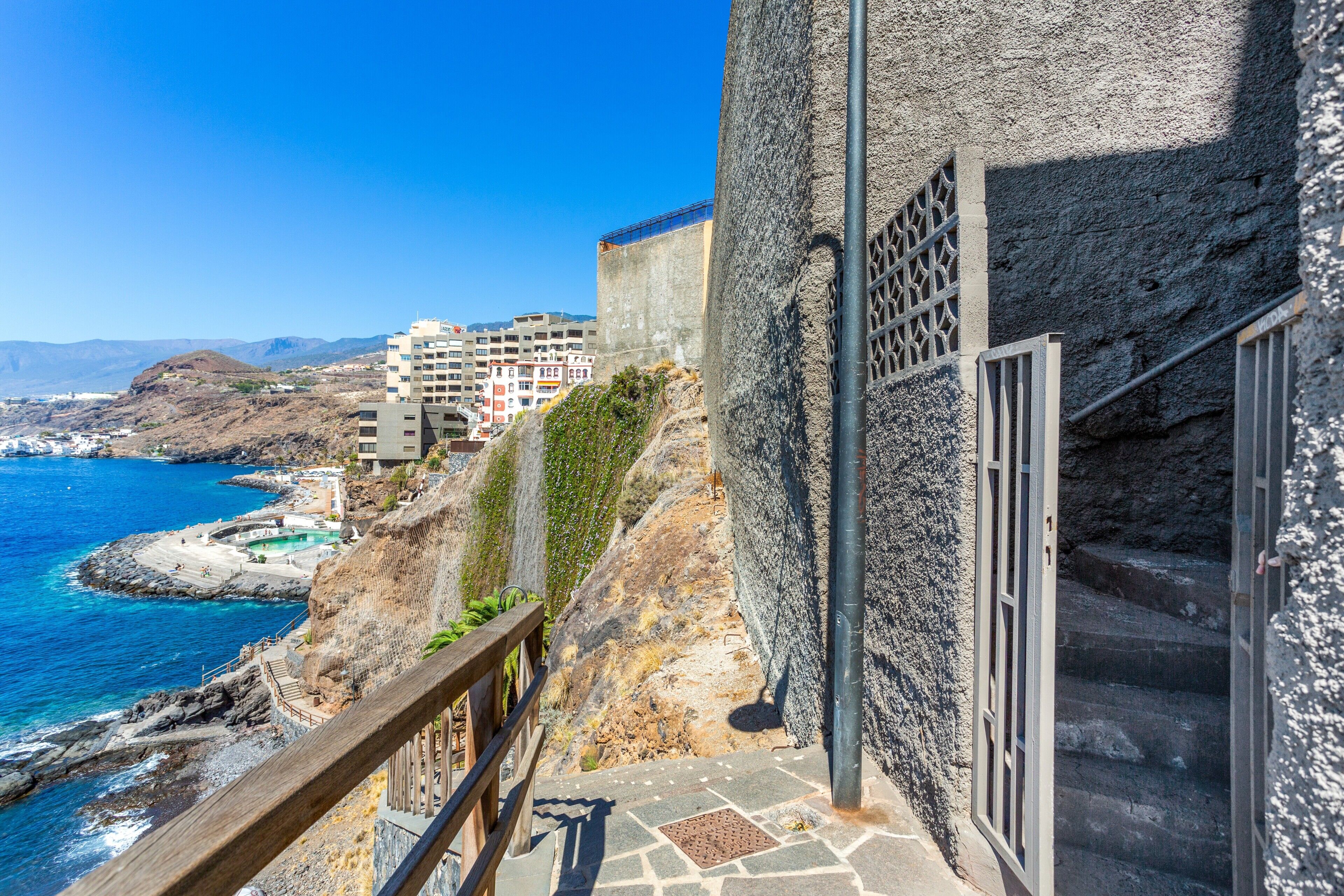 A coastal building with a stone facade, a metal railing, and a view of the sea and mountains.
