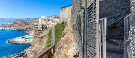 A coastal building with a stone facade, a metal railing, and a view of the sea and mountains.