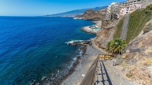 A coastal road with a clear view of the sea, a rocky shoreline, and buildings with balconies overlooking the ocean.
