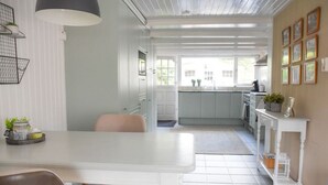 Eine helle Küche mit weißen Schränken, ein Essbereich mit weißem Tisch und Stühlen sowie ein Blick ins Freie durch große Fenster.