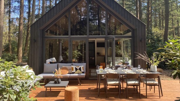 A modern wooden cabin with large windows, a dining area with a table and chairs, and a cozy seating area with a sofa and cushions. - Fantastic luxury forest house | Unwind in nature (Emst)
