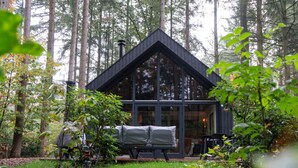 A modern cabin with a triangular roof, large glass windows, and a patio area with a bench and table. - Fantastic luxury forest house | Unwind in nature (Emst)