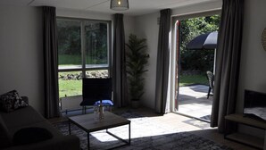 A living room with a sofa, a coffee table, and a TV. There is a large window and a door leading outside. - Comfortable wellness house, everything new, located by the lake. (Ewijk)