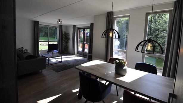 A modern living room with a sofa, a coffee table, and a dining area with a table and chairs. There are large windows providing natural light and views of the outdoors. - Comfortable wellness house, everything new, located by the lake. (Ewijk)