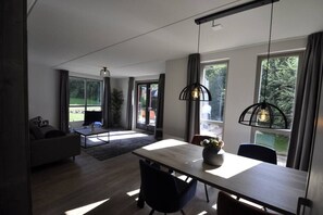 A modern living room with a sofa, a coffee table, and a dining area with a table and chairs. There are large windows providing natural light and views of the outdoors. - Modern wellnesshuis met sauna bij het meer | Ewijk, bij Nijmegen (Ewijk)