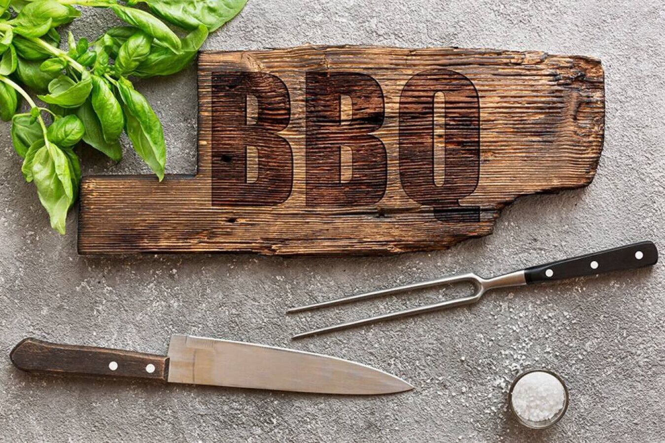 Een rustiek houten bord met daarin het woord 'BBQ' gekerfd, een mes en een kleine schaal met zout op een structuurrijke ondergrond.