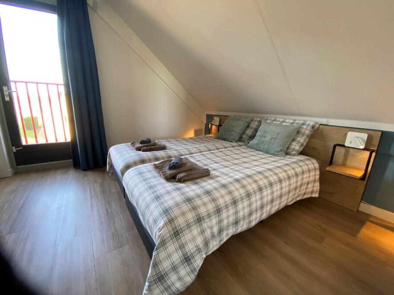 Quarto com teto inclinado, cama com colcha xadrez, duas almofadas e mesa de cabeceira com abajur.