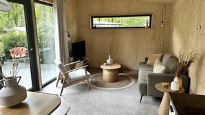A modern living room with a sofa, coffee table, and a TV. There is a window with a view of greenery outside. - VHNT76 · FIKA Luxury holiday home for 4 people at Veluwe (Hoenderloo)