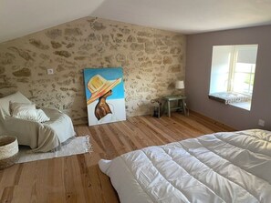 A bedroom with a stone wall, a bed, a sofa, a painting, a lamp, and a window.