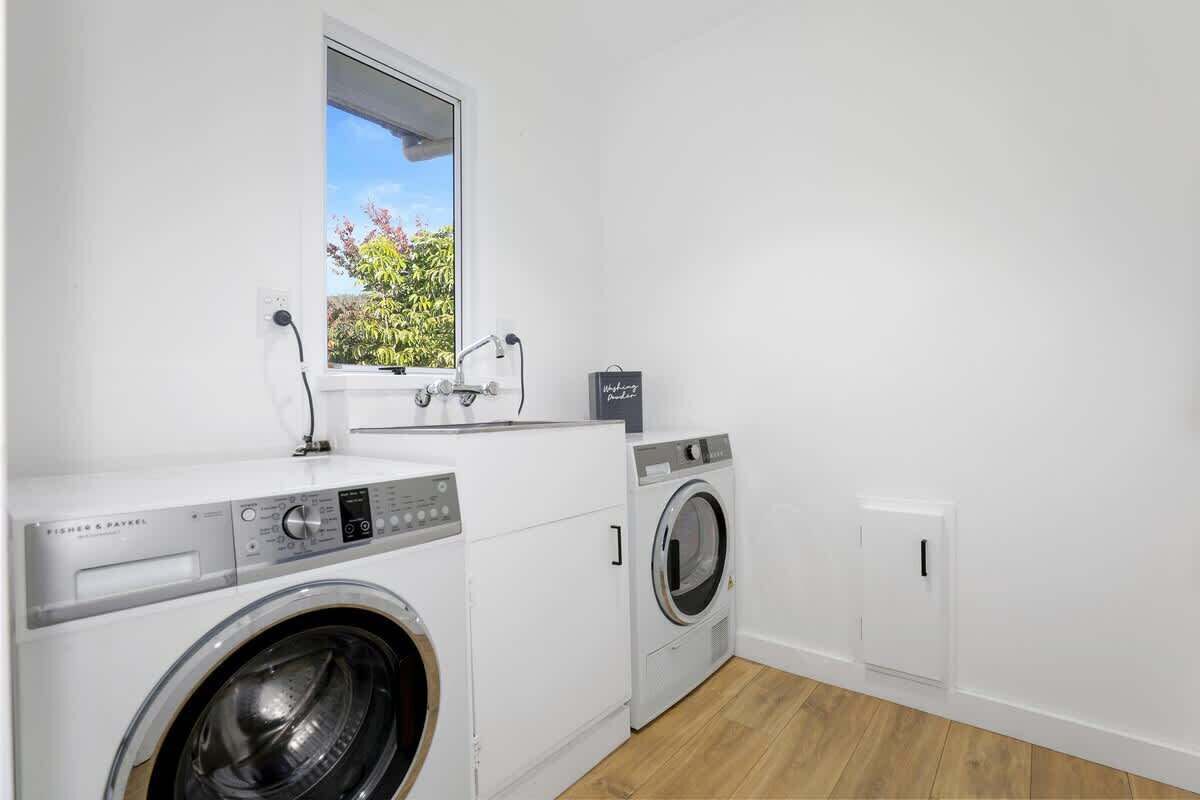 A laundry room with a front-loading washing machine and dryer, a window with a view of greenery, and a small cabinet.