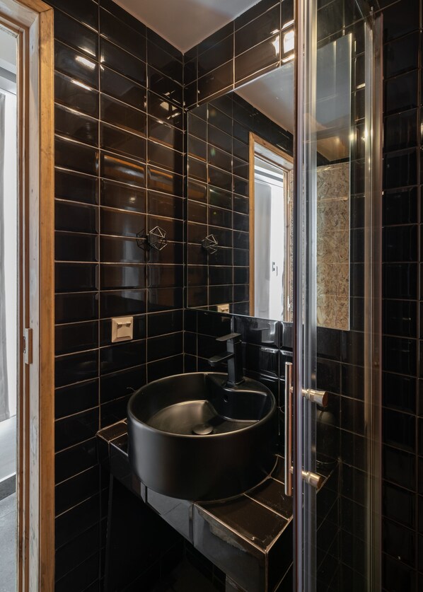 Un baño moderno con paredes de azulejos negros, un lavamanos redondo y una cabina de ducha de vidrio.