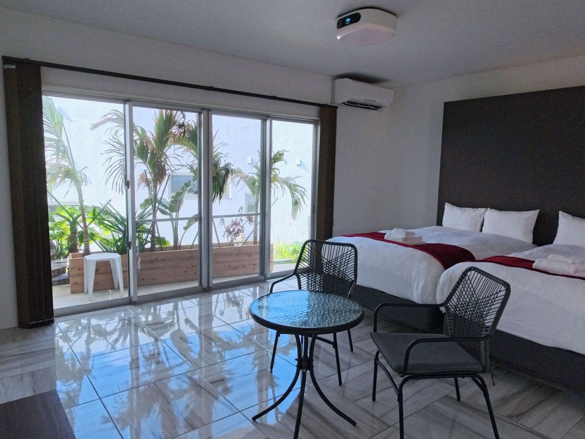 A hotel room with a bed, a table, two chairs, and a view of palm trees through large windows.