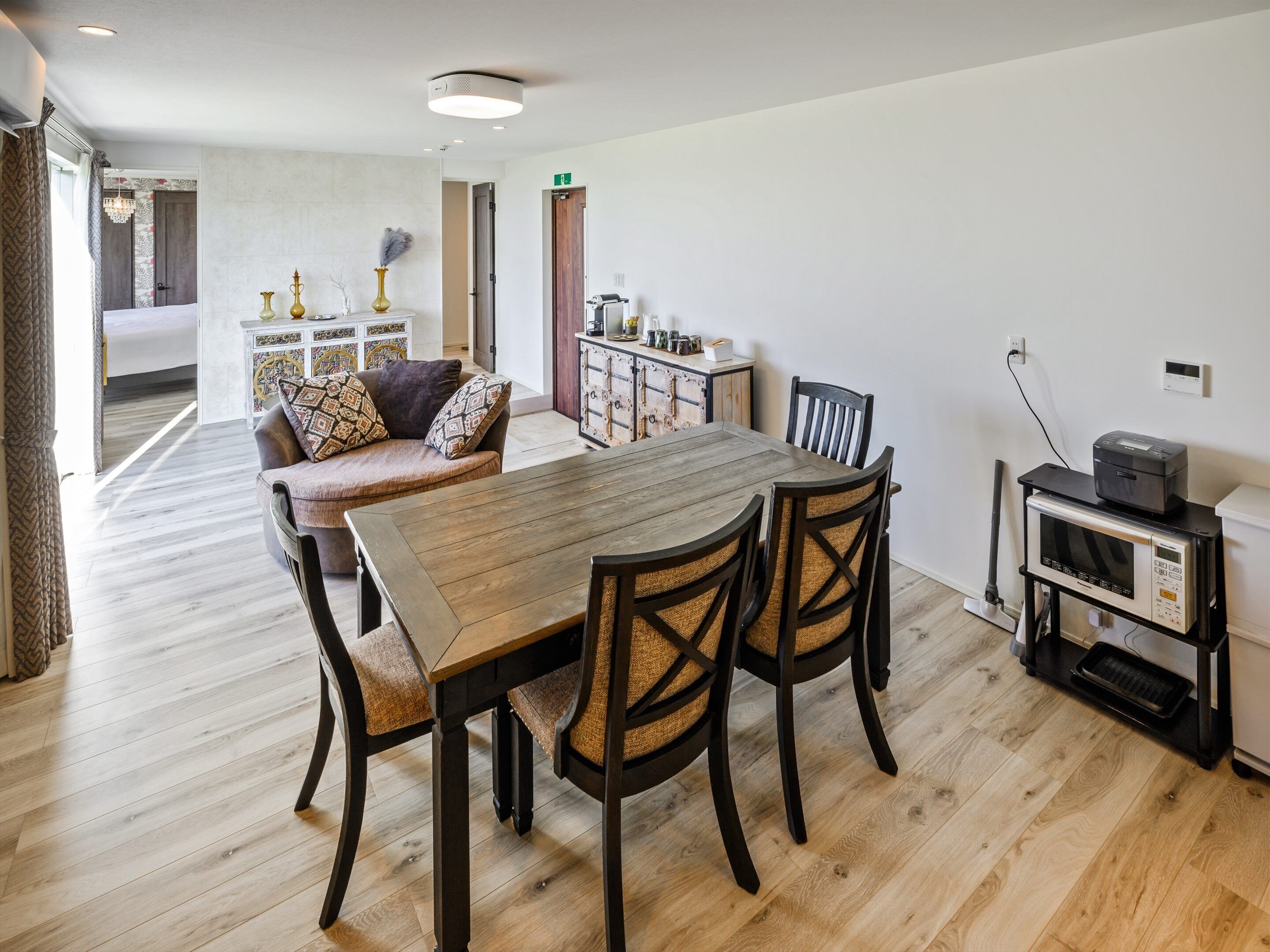 Uma sala de jantar com mesa e cadeiras de madeira, uma cozinha com armários e micro-ondas, e um corredor que dá para um quarto.