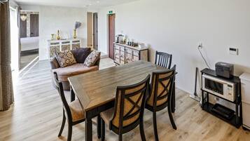 Uma sala de jantar com mesa e cadeiras de madeira, uma cozinha com armários e micro-ondas, e um corredor que dá para um quarto.
