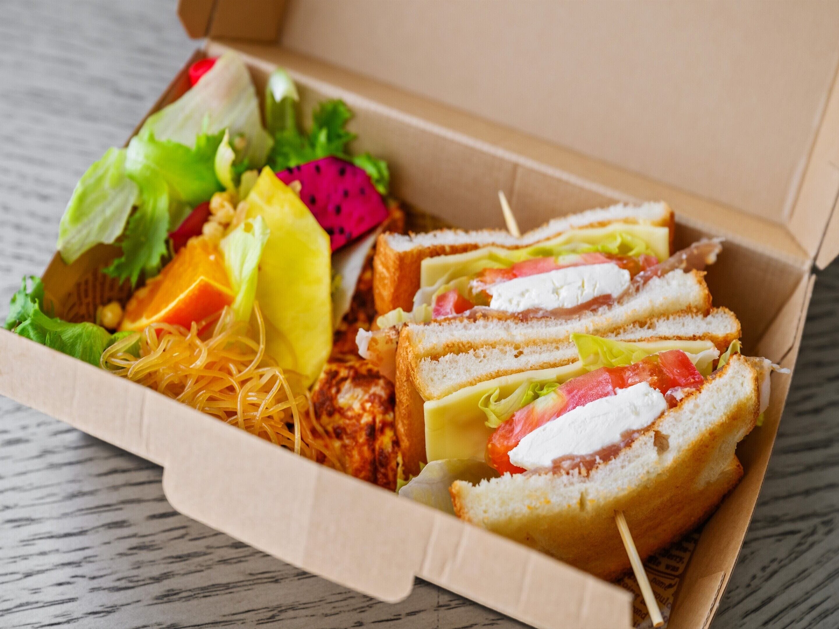 Una caja de cartón con un sándwich de lechuga, tomate y queso, y una ensalada acompañante de lechuga, zanahorias y pitahaya.