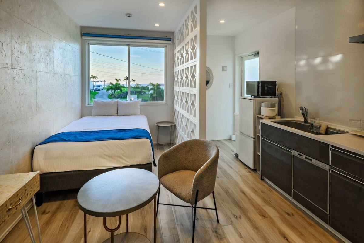 Quarto de hotel moderno com uma cama, uma mesinha redonda, uma cadeira, cozinha compacta e vista para o exterior.