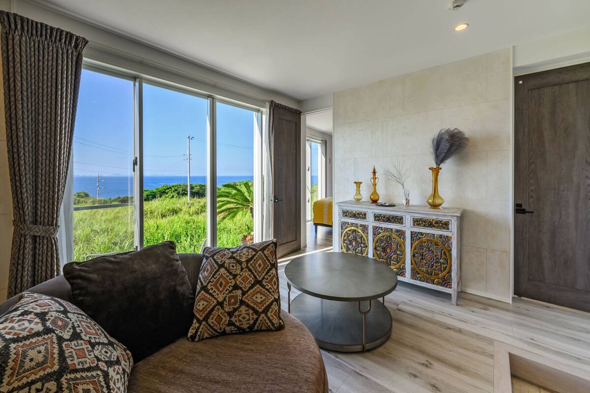 Uma sala de estar espaçosa com uma grande janela que oferece vista para o oceano, uma mesa de centro redonda e um armário branco com objetos decorativos.